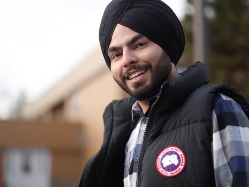 A bearded man wearing a black turban, flannel shirt, and puffer vest, smiling toward the camera