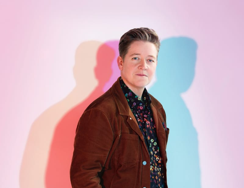 Ivan Coyote posing in front of a white wall cast in blue and pink light