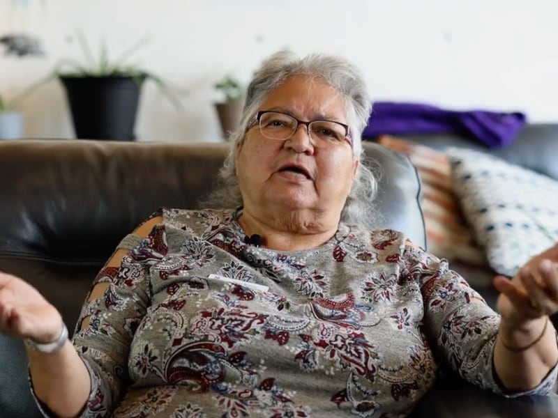 A woman wearing glasses and patterned shirt gestures towards the camera