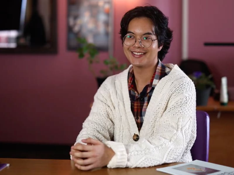 A man wearing glasses and a white sweater sits at a table with hands folded