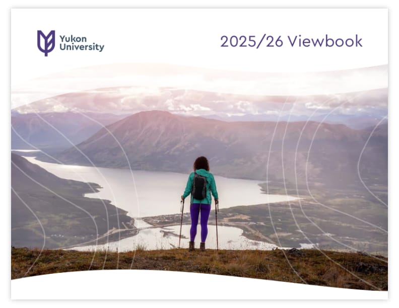 A woman wearing a teal jacket and purple pants, holding hiking poles, standing overlooking a lake and mountains