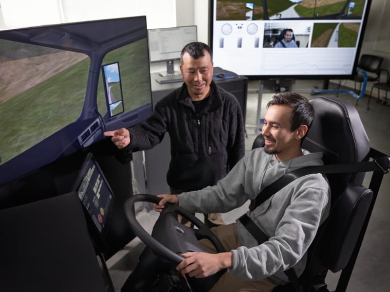 A smiling student operates a driving simulator while an instructor points to the screen