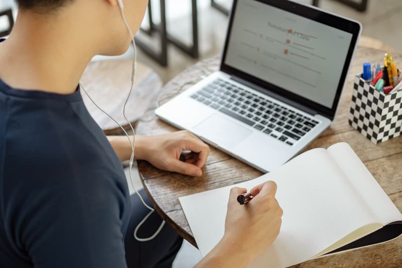 A person with short hair wearing wired earbud headphones looks at an open laptop and holds a pen ready to write in an open notepad.