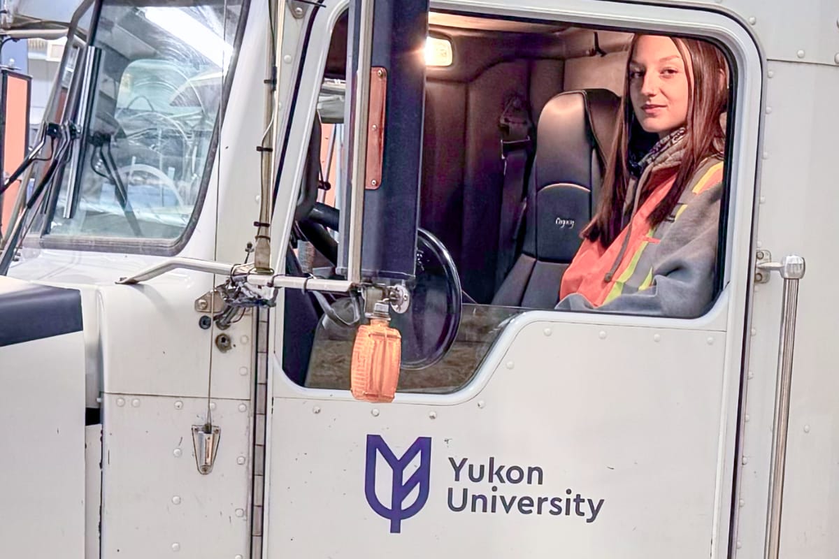 A woman with long hair wearing a safety vest and sitting in the driver's seat of a large truck with a Yukon University logo on the door