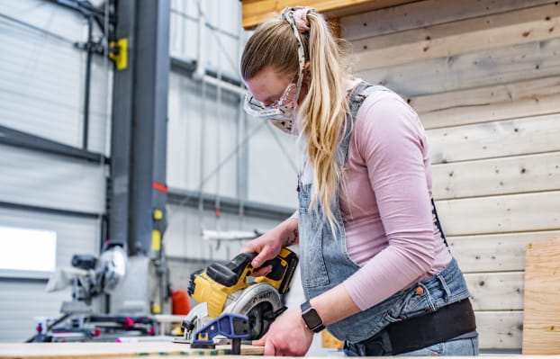 A woman with a ponytail and wearing overalls cutting a sheet of plywood with a circular saw