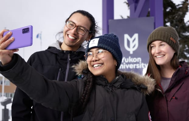 Three smiling students taking a selfie photo together in front of a Yukon University sign
