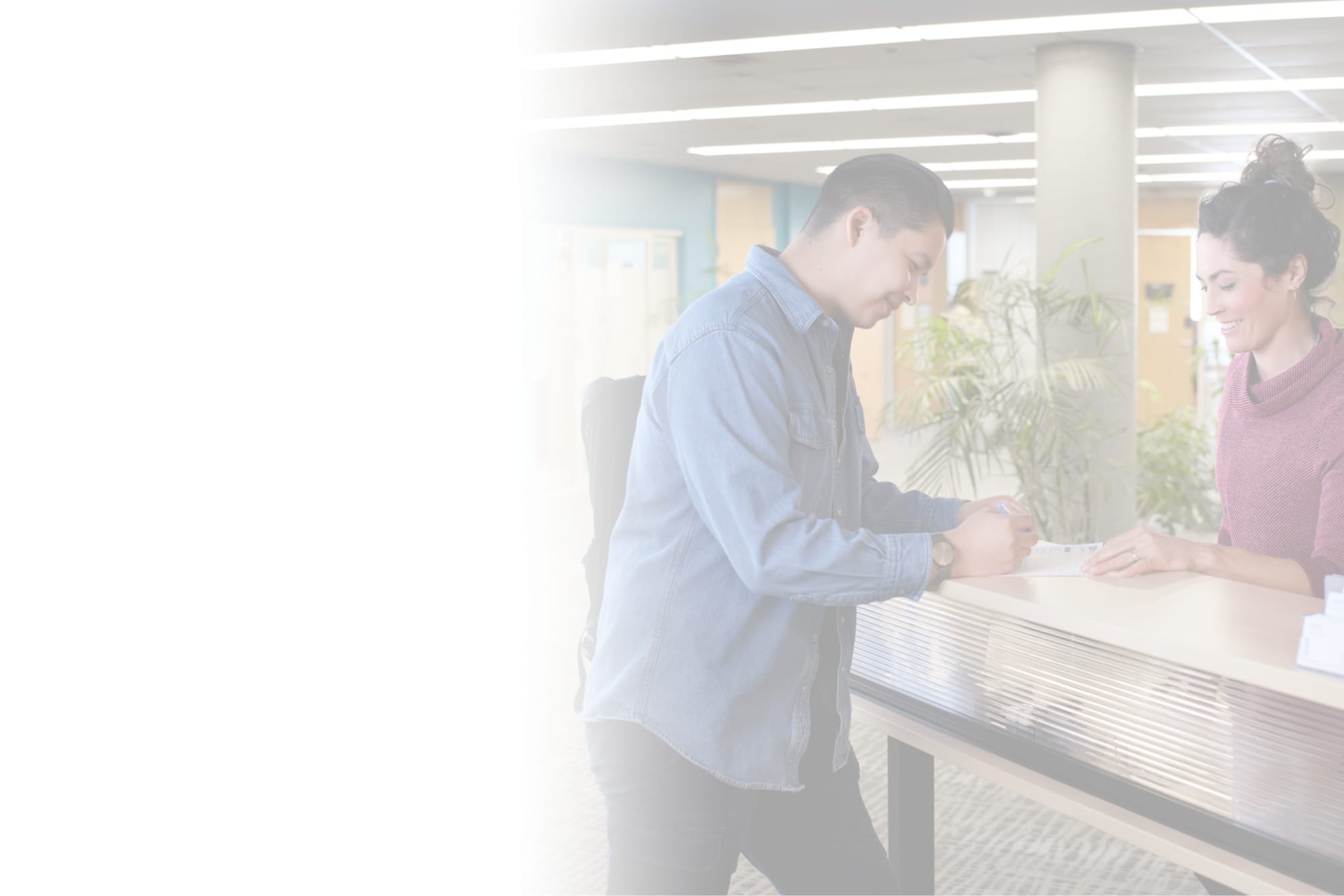 A student filling out a paper form while a smiling staff member looks on behind a desk