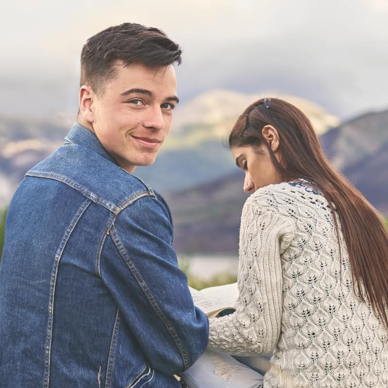 Two students sitting outside with a mountain view. One of them looking back toward the camera.