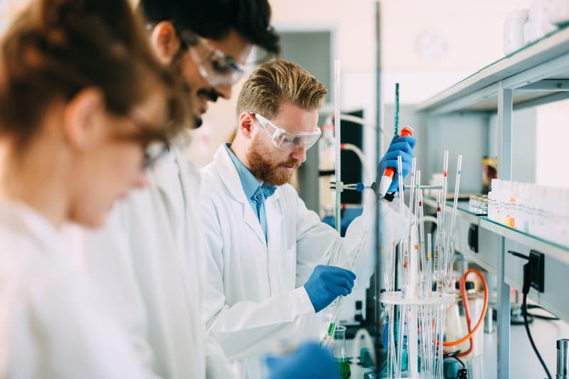 Three people at a lab bench wearing lab coats and safety glasses, using micropipettes.