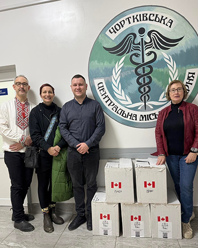 Two men and two women stand on either side of a pile of white cardboard boxes with small Canadian flag stickers. They pose standing against a white wall with a large painted Ukrainian hospital logo. The caption reads "Chortkiv Central City Hospital Receiving Medical Supplies from UCAY"