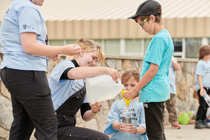 A kids camp counsellor pours a clear liquid through a funnel with the help of two young kids