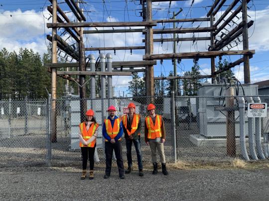 Four researchers in reflective vests and hard hats stand in front of a power line