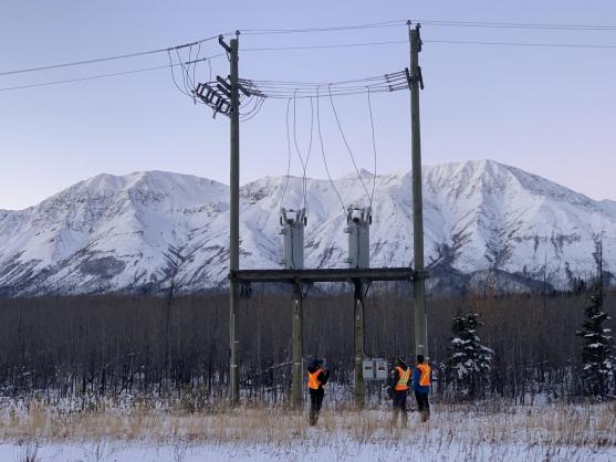 Three researchers in reflective vests look up at a power line in front of a mountain range