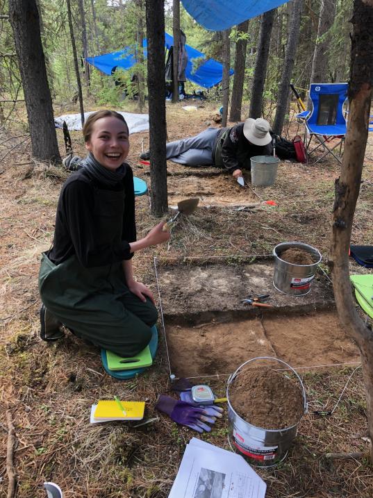 A smiling student researcher poses with a shovel at an archeological dig site