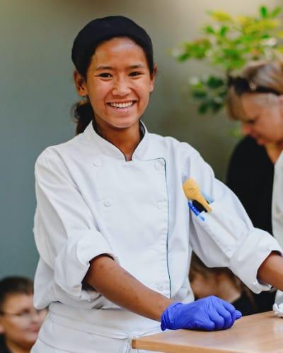 Culinary arts student wearing a white apron and purple gloves, smiling toward the camera