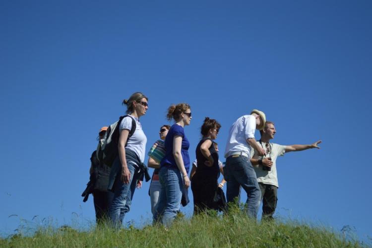 A group of people looking out over a hill. The leader is pointing into the distance while reaching for binoculars that are hanging on his neck from a strap.