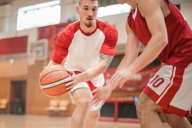 A bearded man in a white shirt dribbles a basketball around a man in a red shirt