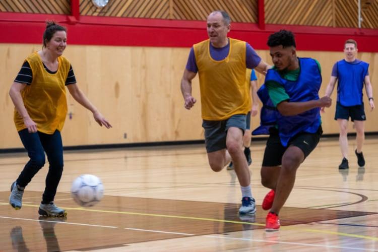 A male student wearing a blue jersey kicks a soccer ball in front of two faculty members wearing yellow jerseys