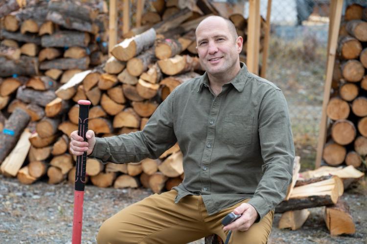 Stephen Biggin-Pound sits on a stump in front of piles of split and stacked firewood