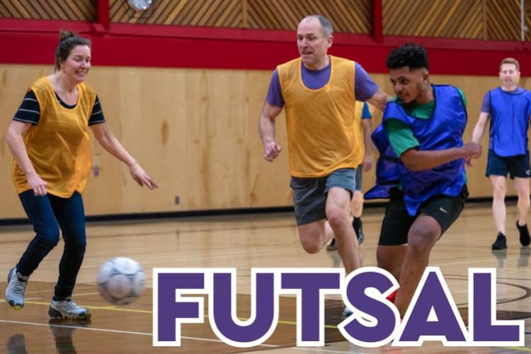 A student in a blue jersey kicking a soccer ball while two staff members in yellow jerseys pursue the ball