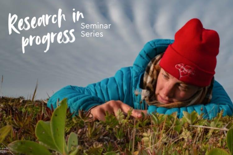 A woman wearing a red toque and blue puffy jacket lays down among some short green plants.
