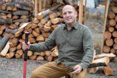 Stephen Biggin-Pound sits on a stump in front of piles of split and stacked firewood