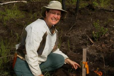 A woman wearing a bucket hat crouching in a forest and pointing to a stake in the ground with an orange ribbon tied to it