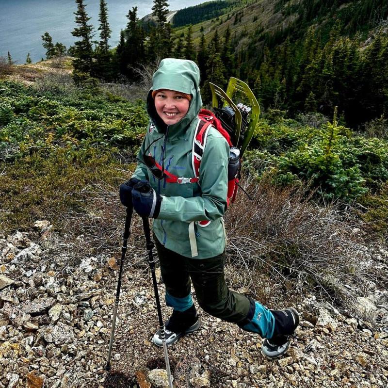 A person wearing a hooded jacket and a backpack, holding hiking poles, posing on a hiking trail above a river with mountains in the background