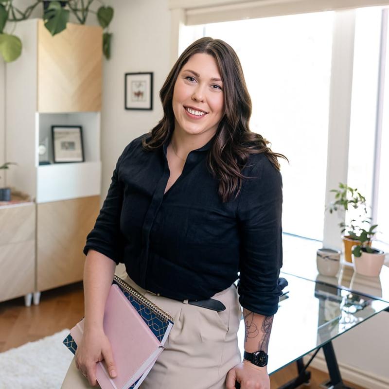 A professional woman in a black shirt holding two notebooks, leaning against a glass desk